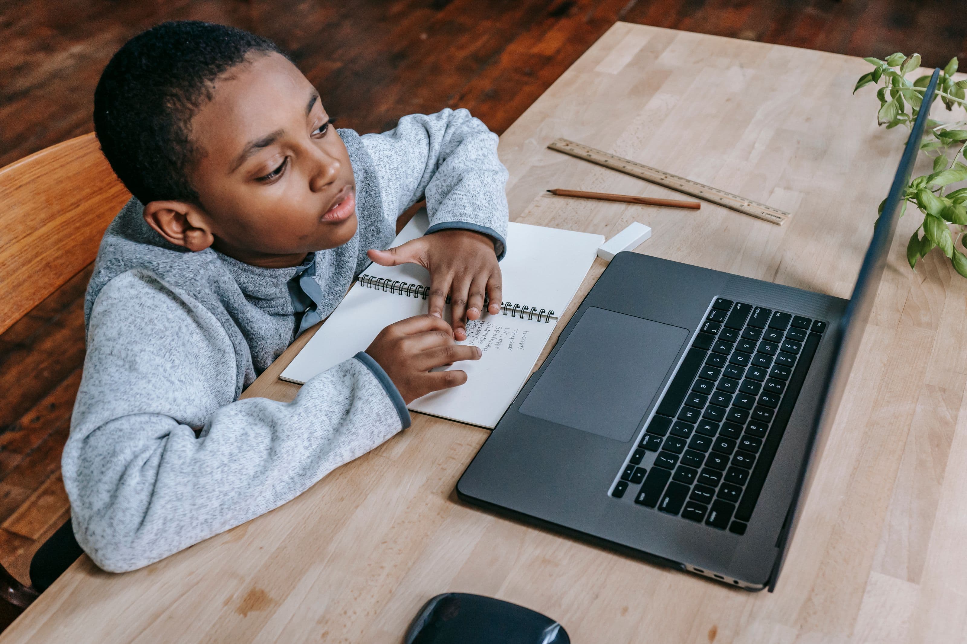 A boy writes in a notebook beside an open laptop at a wooden table indoors