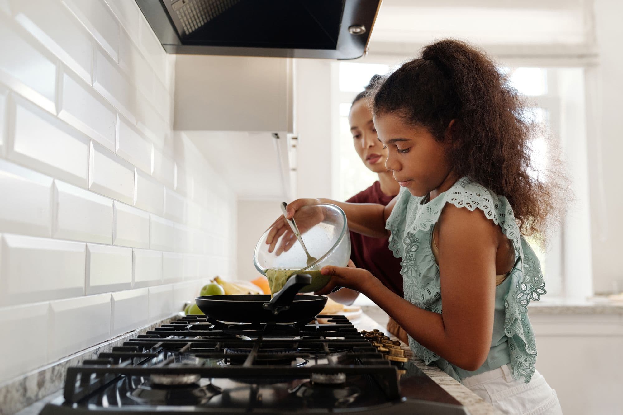 A girl tips batter from a bowl into a frying pan while an adult steadies the hob
