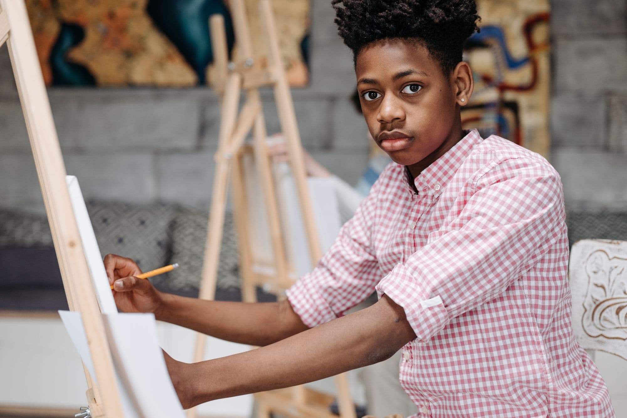 A teenage boy sketches at a wooden easel in a room hung with bright artwork