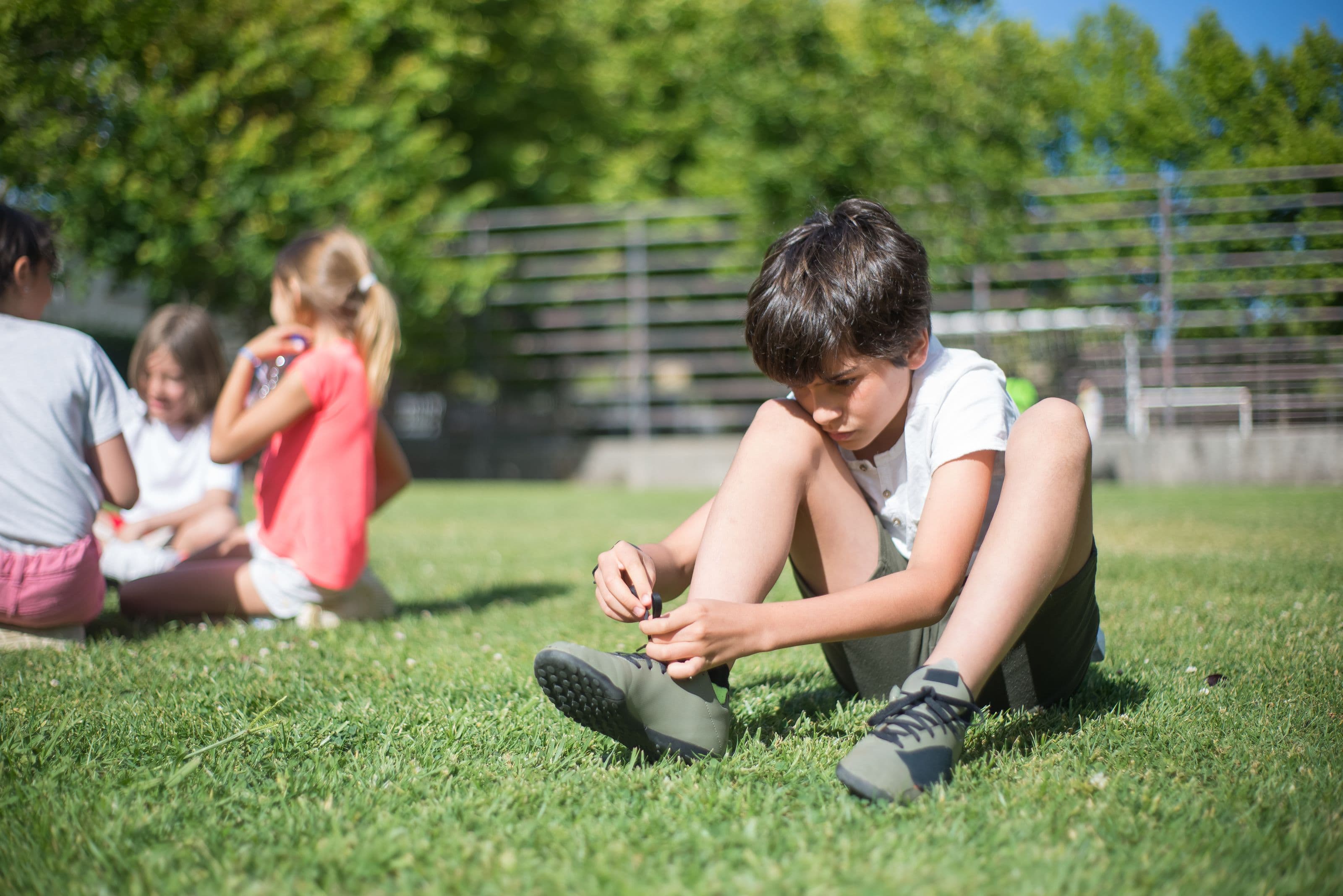 A boy sits on the grass tying his own shoelace while other children rest nearby