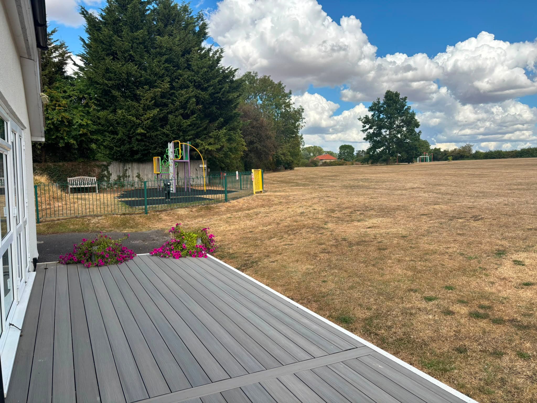 A decked terrace with flower planters looks out over a fenced play area and an open playing field beyond