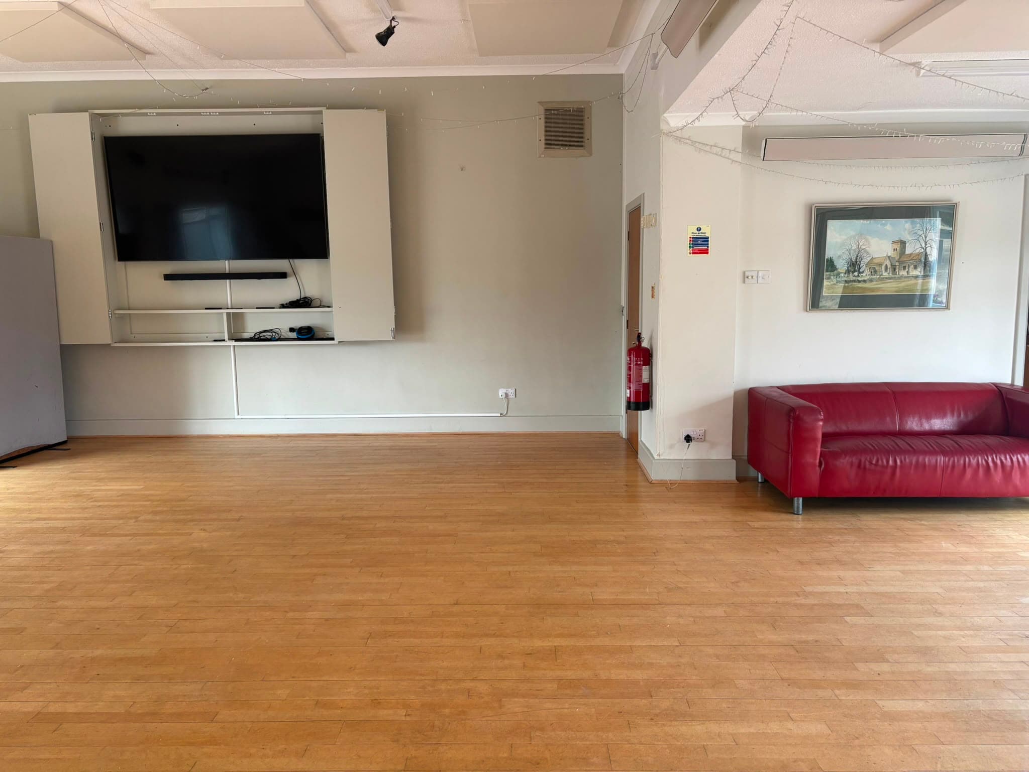 A large hall with a wooden floor, wall-mounted screen and a sofa along one wall