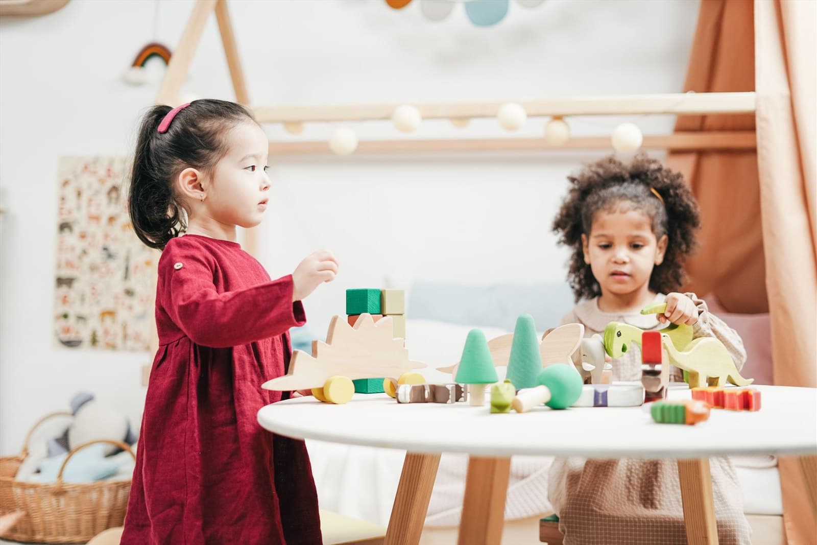 Two young girls arrange wooden blocks and carved animals on a round table in a calm playroom