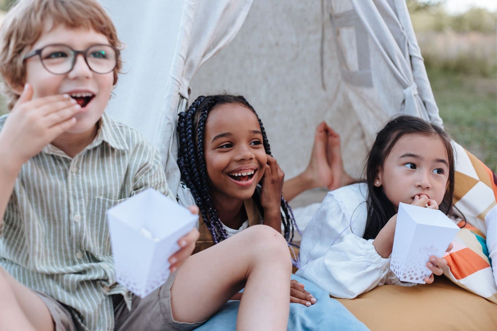 Three children sit together outside a canvas tent, laughing and sharing snacks in late afternoon light