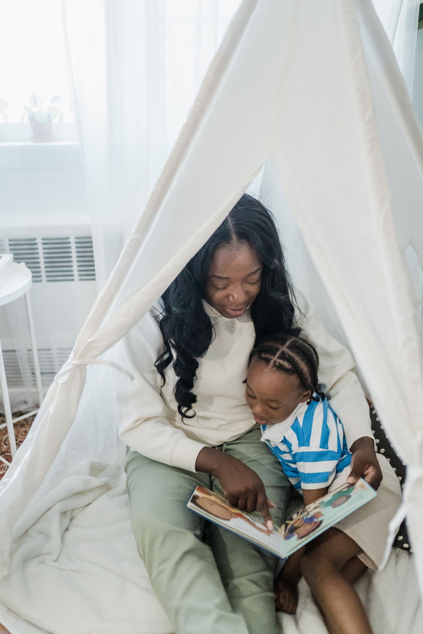 A woman and her young daughter sit inside a fabric play tent at home, reading together