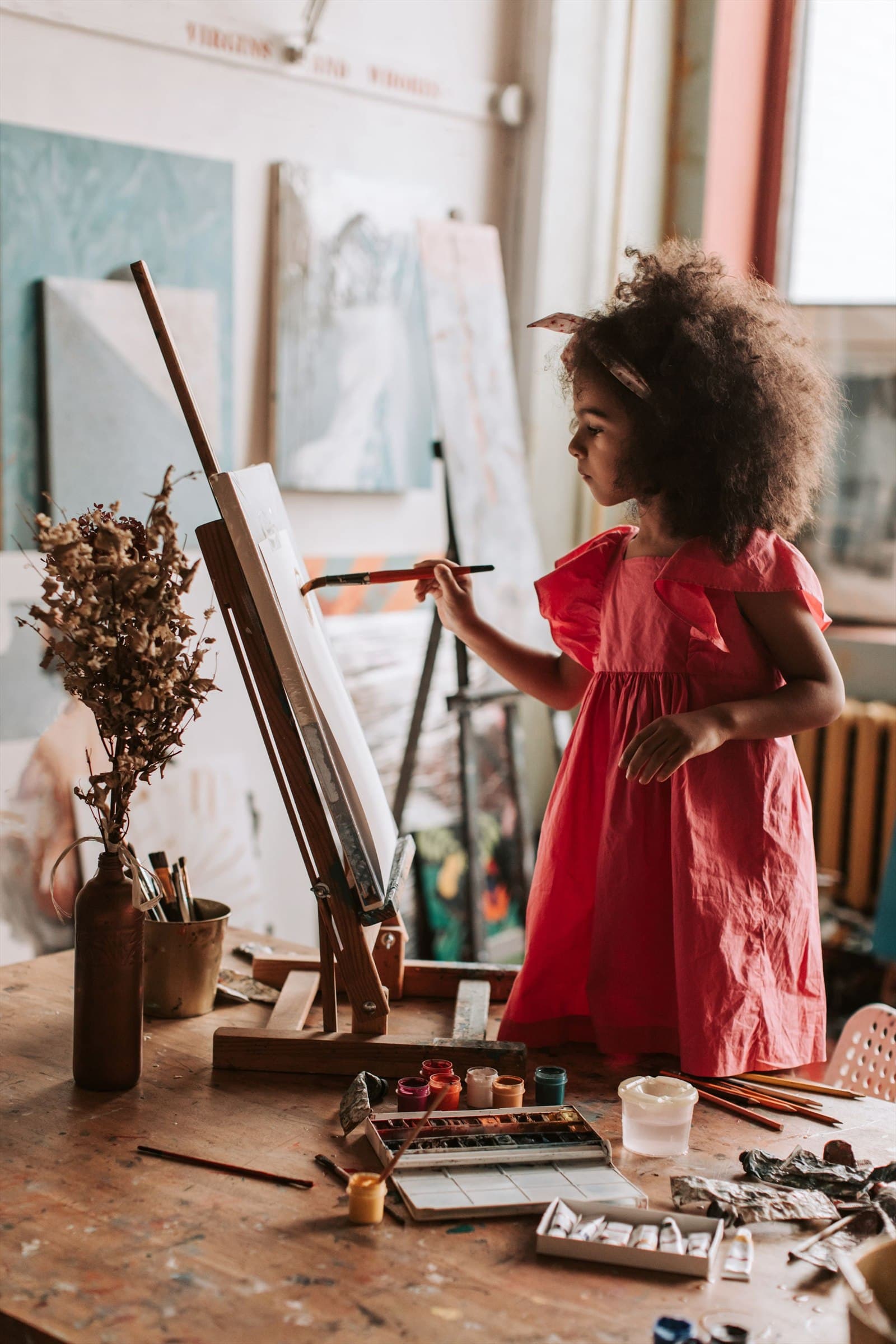 A girl in a red dress paints at a wooden easel in a light-filled studio