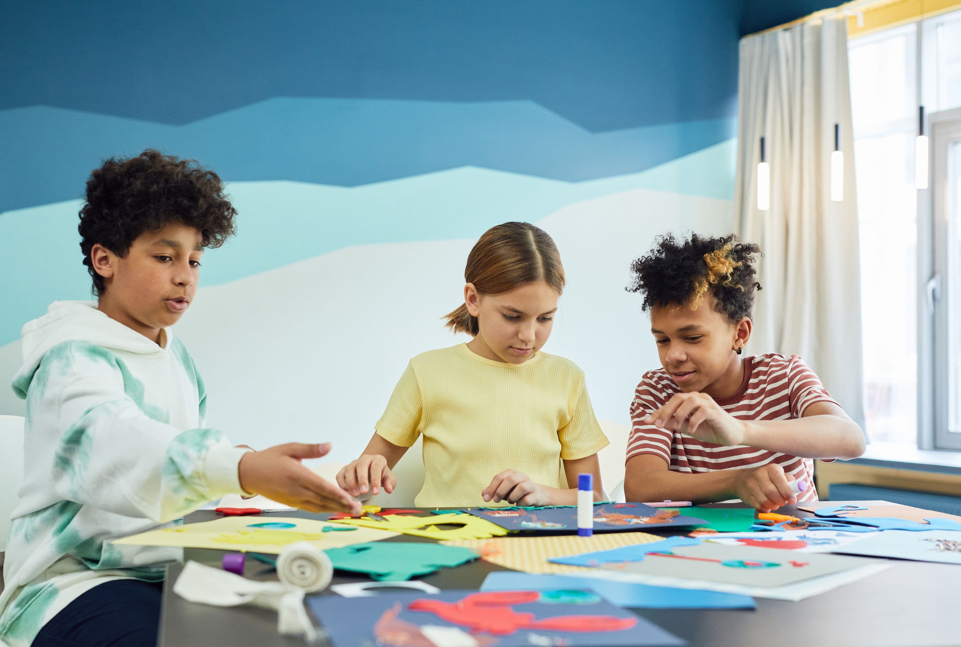 Three young people cut and glue coloured paper at a table beside a bright window