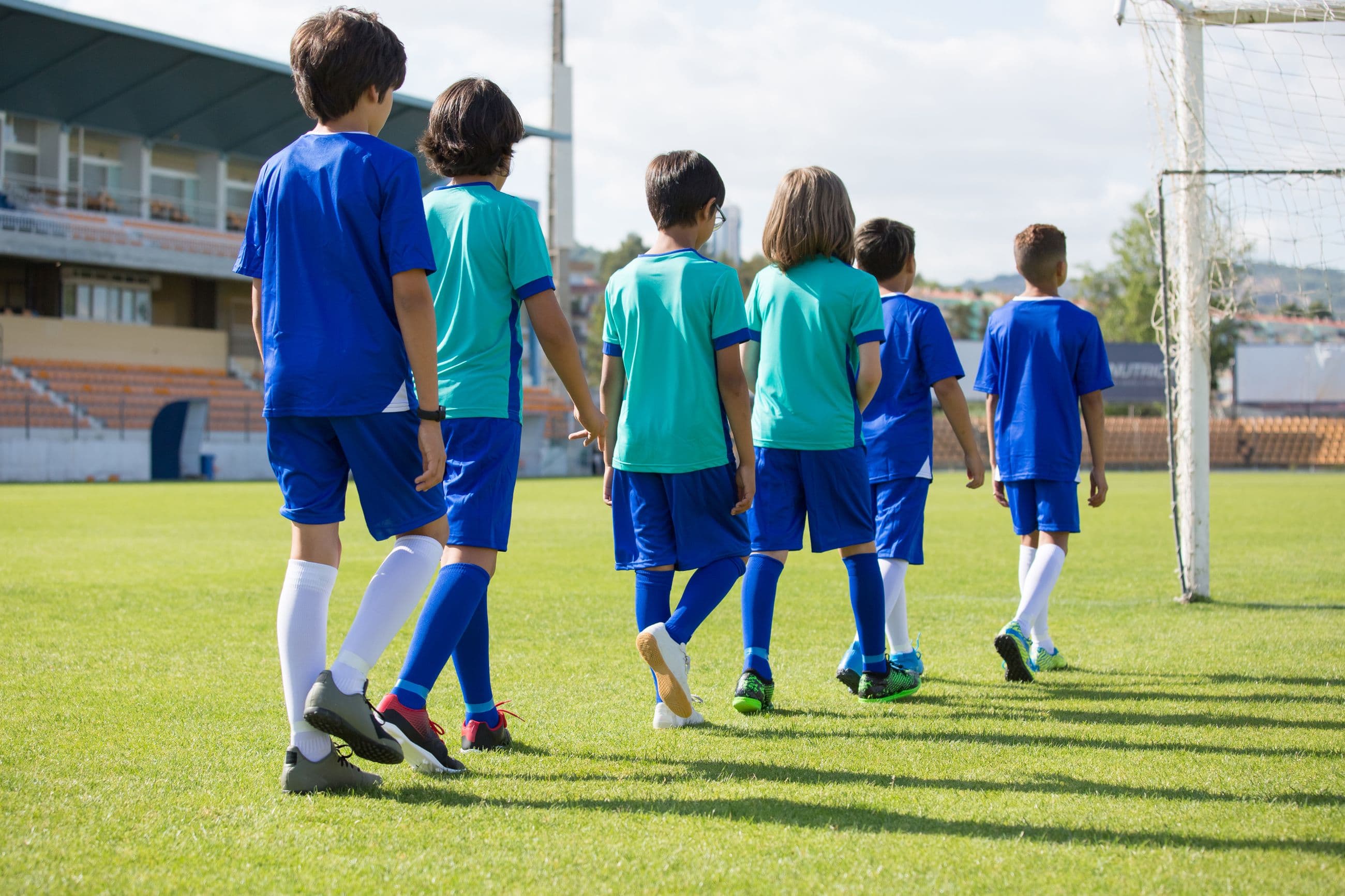 Children in football kit walk together towards the goal across a green grass pitch
