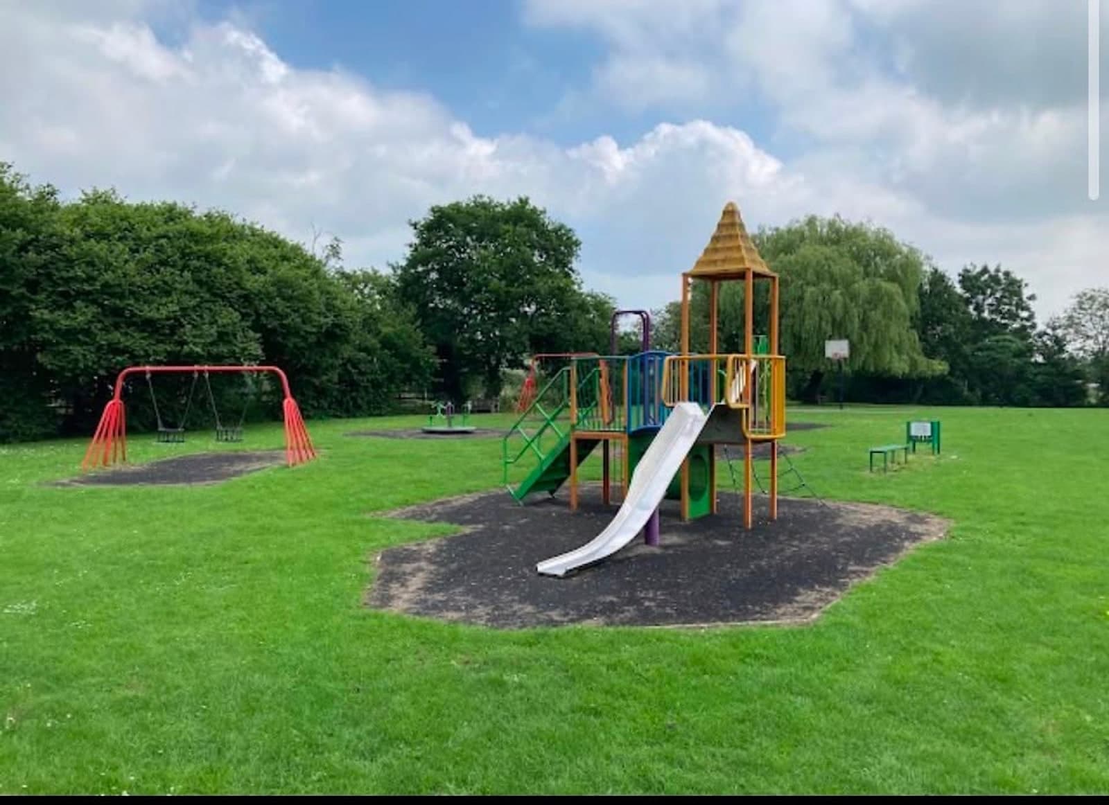 A play tower with a slide, swings and a basketball hoop set around a wide mown green